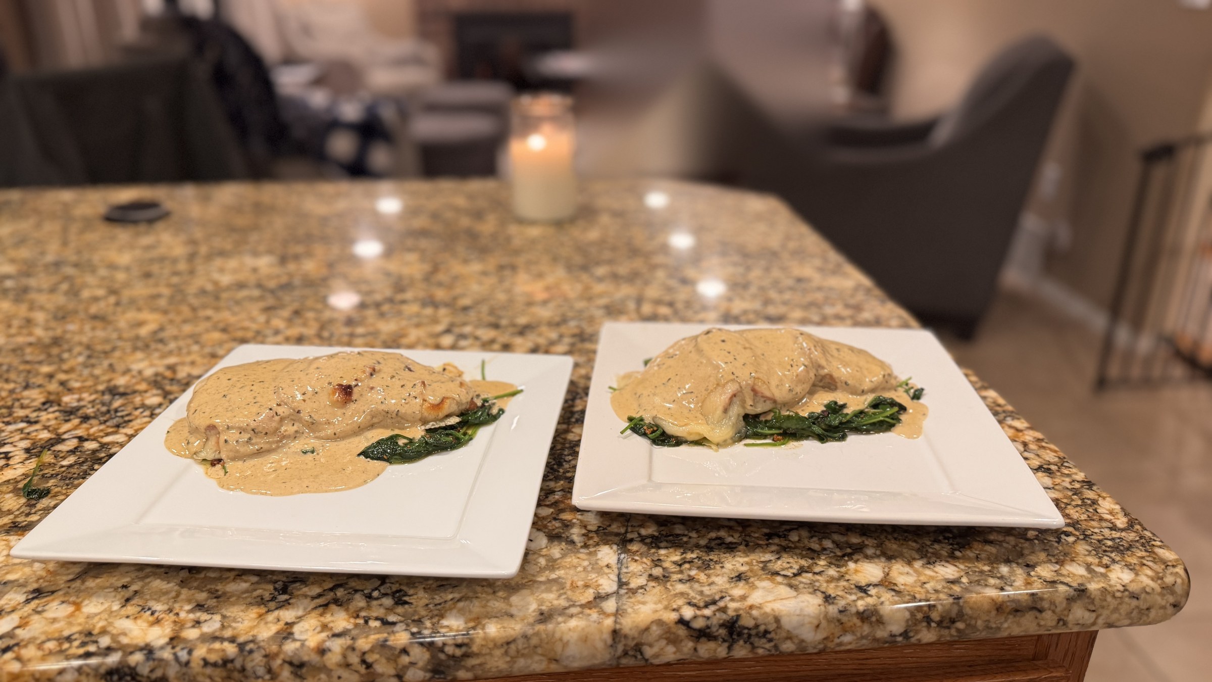 Two white square plates of Chicken Dijon on a granite countertop: each pan-seared chicken breast smothered in creamy Dijon sauce flecked with pepper, set on a bed of wilted spinach, with a lit candle in the softly blurred background